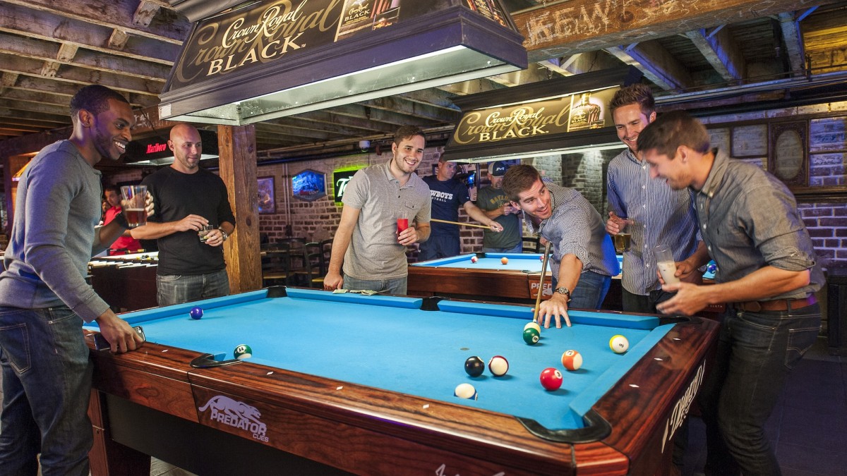 Group of friends playing pool inside a lively Savannah bar, gathered around a blue felt pool table with drinks in hand beneath exposed wooden beams.