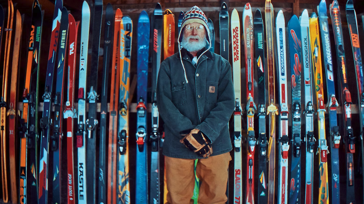 Man standing in front of a colorful wall of vintage skis, wearing a winter jacket, knit hat and gloves inside a rustic indoor setting.