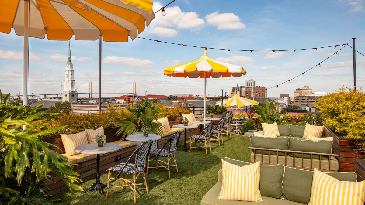 Outdoor rooftop lounge at Perry Lane Hotel’s Peregrin Rooftop Bar, featuring green seating, yellow-and-white umbrellas, café tables and sweeping views of the Savannah skyline and church steeple under a blue sky.