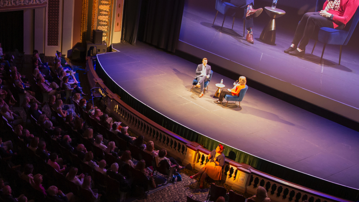Wide view of a theater audience watching an onstage conversation between two speakers seated in chairs beneath a large screen.