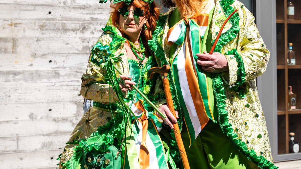 Two people dressed in festive green St. Patrick’s Day attire, wearing shamrock-themed outfits and accessories, pose together on a brick street in Savannah.