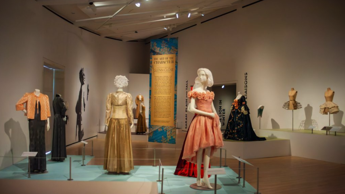 Gallery view of a museum exhibition displaying period costumes on mannequins, with dresses arranged on platforms and a central banner highlighting the Glenn Close costume collection.