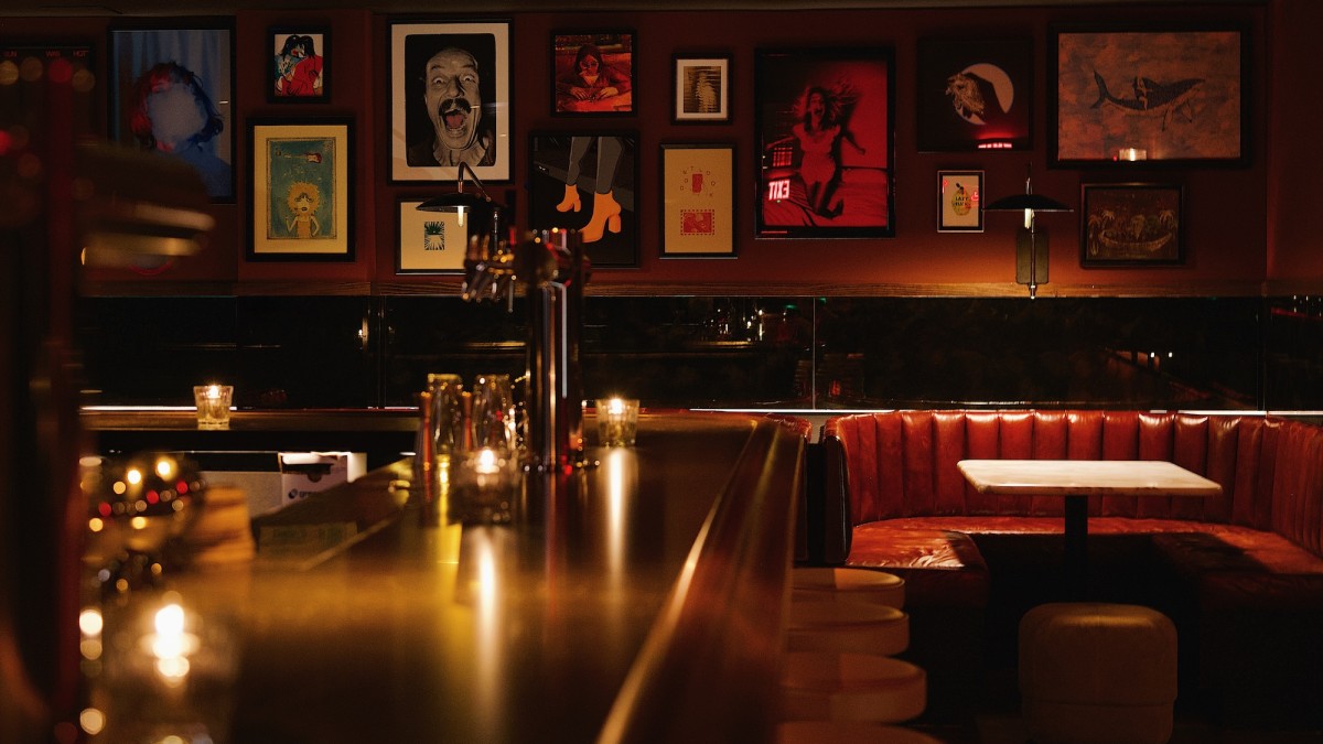 Dimly lit speakeasy-style lounge with red leather banquettes, candlelit tables, framed art-lined walls, and an intimate bar setting.