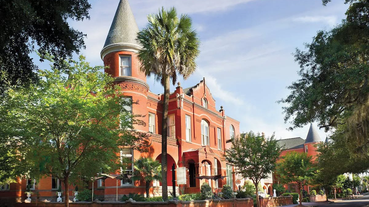 Exterior of Hotel Bardo Savannah, a red-brick historic building with a turreted corner, arched windows, palm trees and a tree-lined street in the foreground.