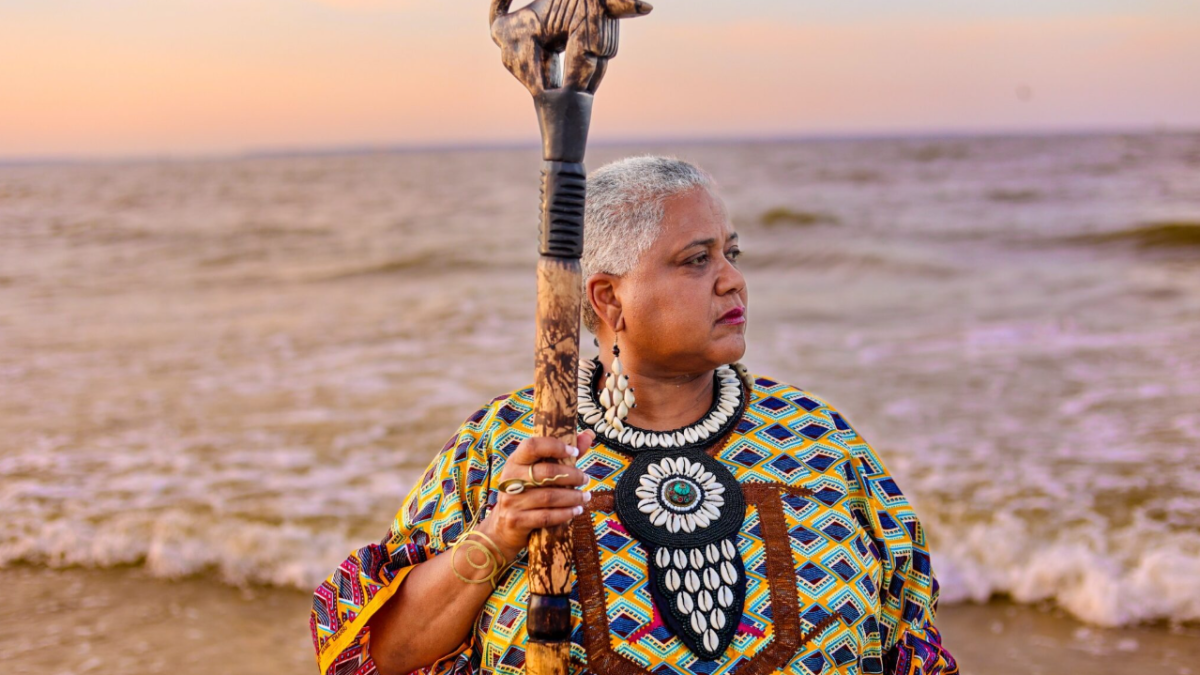 A woman dressed in African attire holding a staff while looking out onto the ocean.