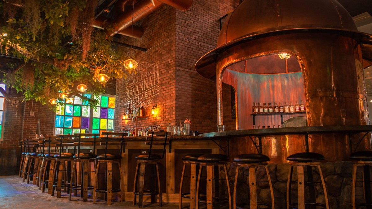 Interior of Old Savannah Distillery with copper still, bar seating, exposed brick and warm lighting.