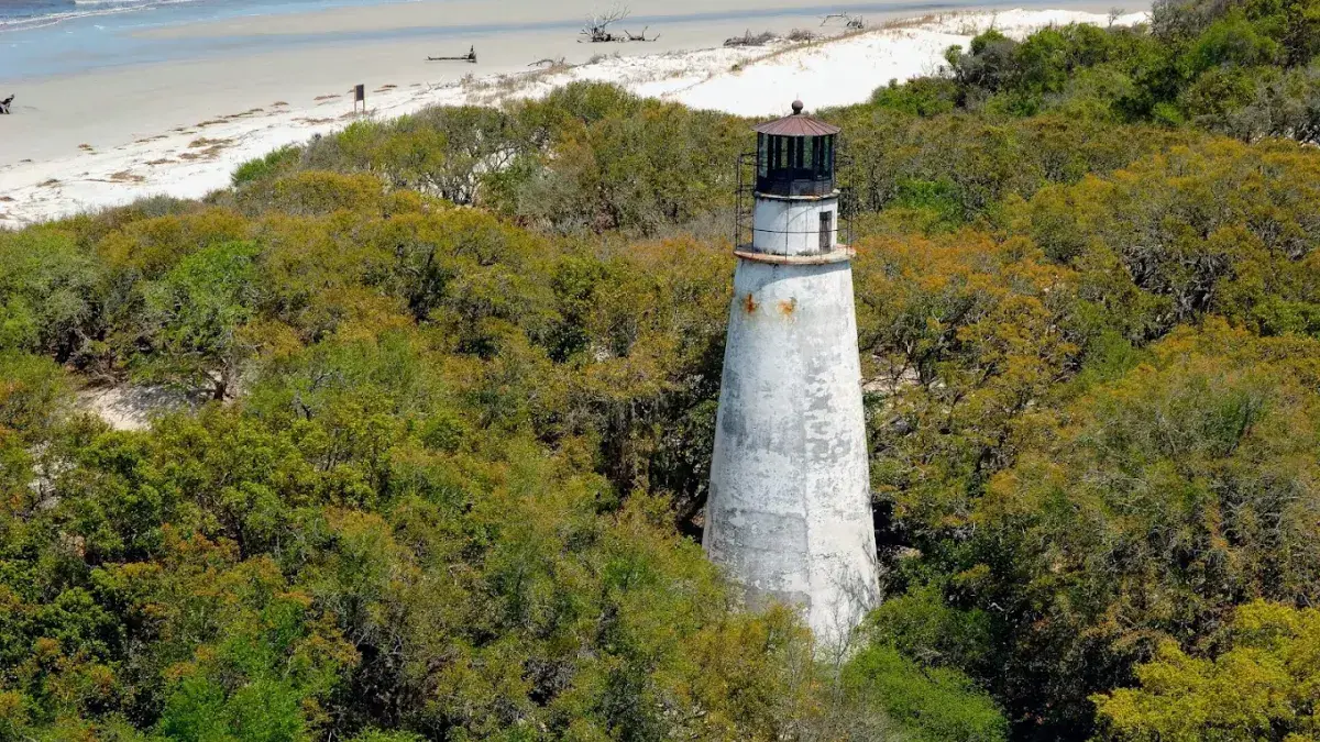Historic white lighthouse rising above dense coastal forest near a sandy shoreline, with ocean waves visible in the background.