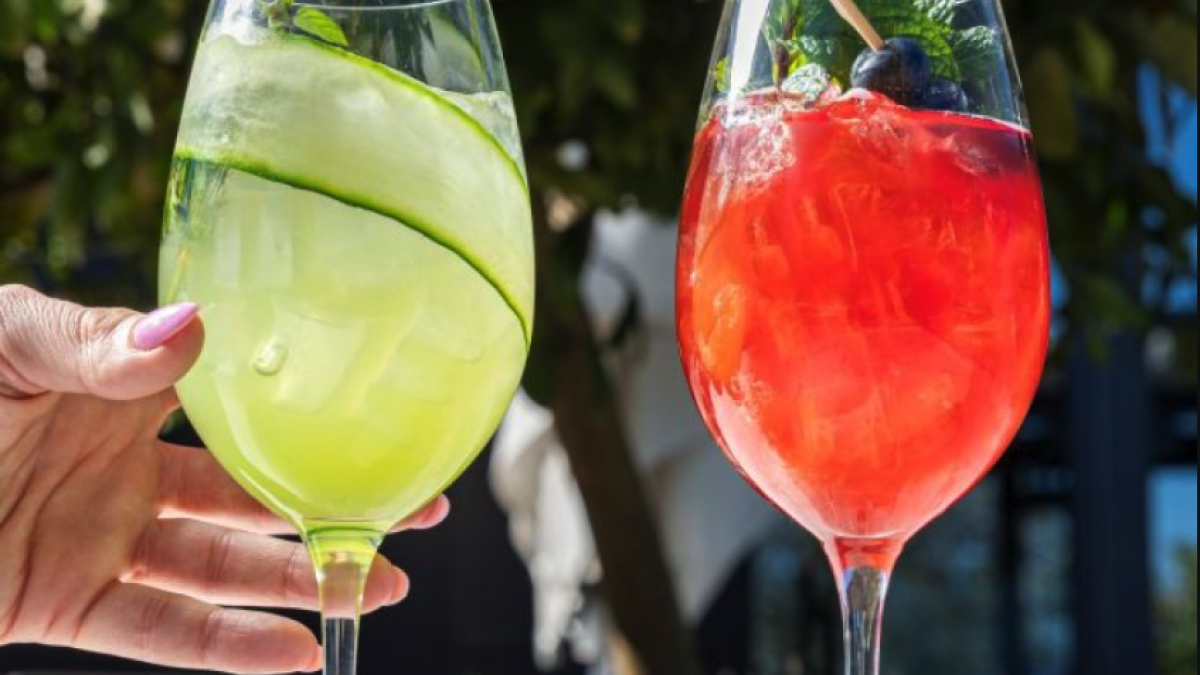 Two colorful brunch cocktails—one green with a cucumber garnish and one red with berries and mint—held outdoors in a sunny garden setting.