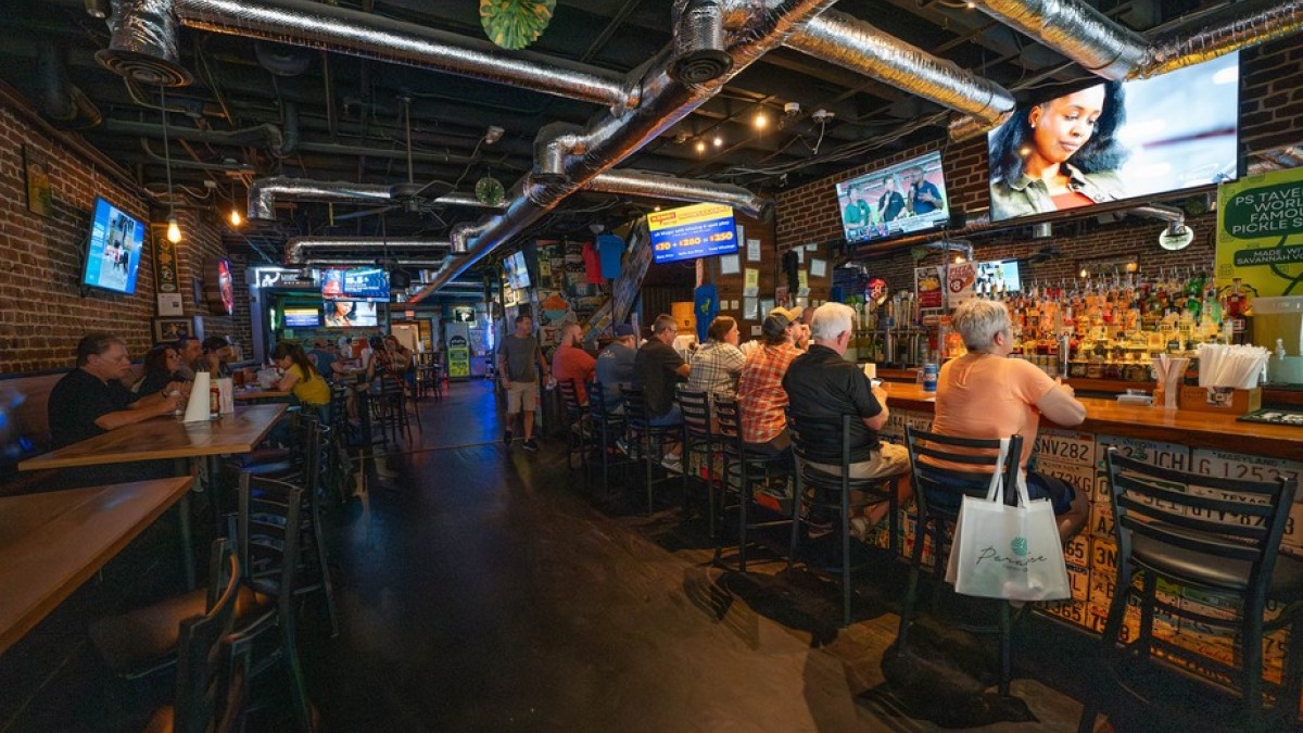 A lively interior view of PS Tavern shows guests seated at a long bar watching sports on multiple TVs, with exposed brick walls and a warm, bustling atmosphere.