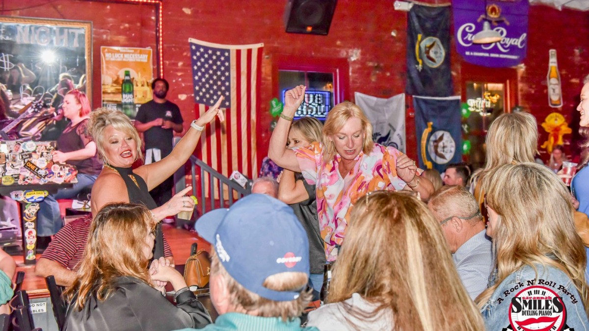 A lively crowd gathers inside Savannah Smiles Dueling Pianos as guests dance, sing along and enjoy live music in a vibrant bar setting.