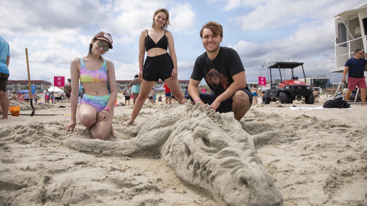 Three people pose on the beach beside a detailed sand sculpture during the SCAD Sand Arts Festival on Tybee Island.