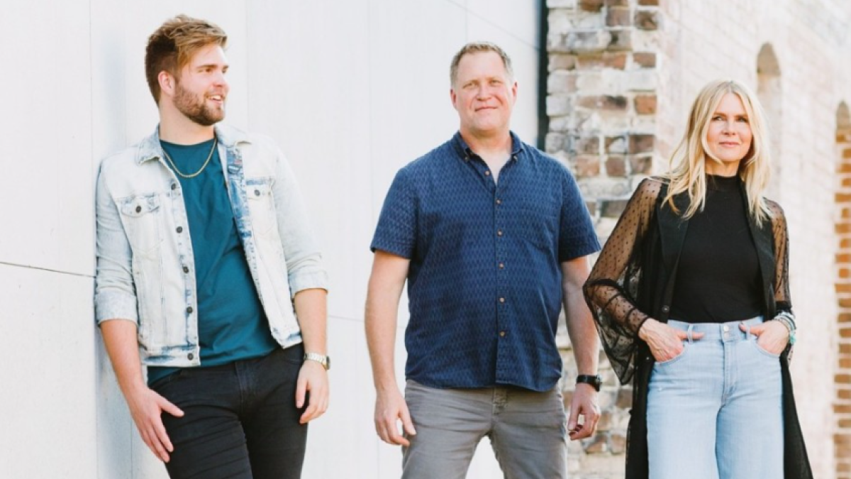 Three members of Scarlet Trio walk side by side outdoors along a brick and stucco wall.