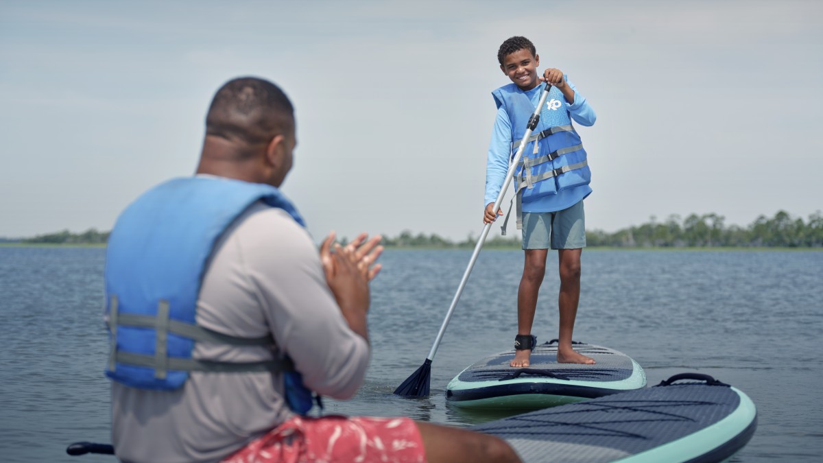 A child stands on a paddleboard smiling while an adult sits nearby, both wearing life vests on calm coastal waters near Tybee Island.