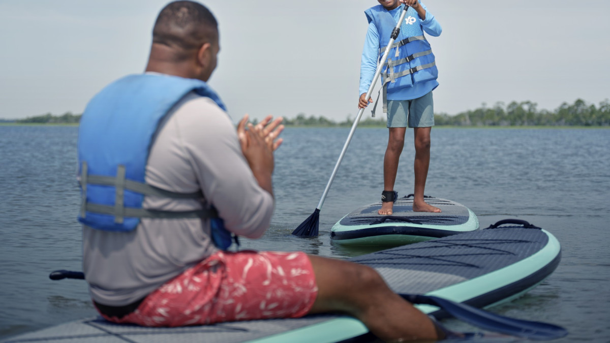 A child stands on a paddleboard smiling while an adult sits nearby, both wearing life vests on calm coastal waters near Tybee Island.