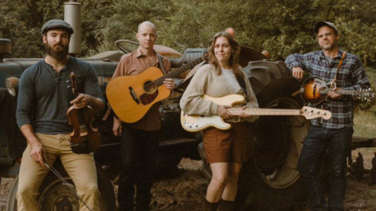Members of the band Swamptooth pose outdoors with string instruments in front of a tractor.
