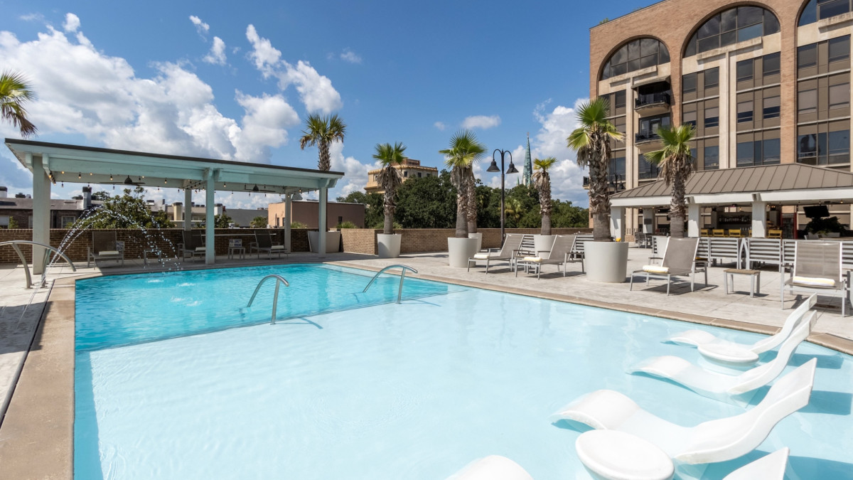 Outdoor pool deck at The DeSoto Savannah featuring a shallow sun shelf with in-water lounge chairs, palm trees, poolside seating and views of the hotel's rooftop terrace beneath a bright blue sky.