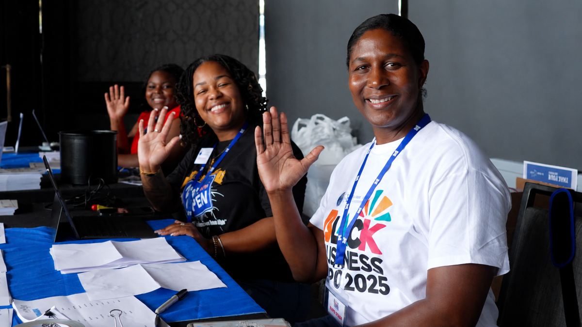 Three volunteers smile and wave while seated at the registration table during the Savannah Black Business Expo. Event materials, laptops and check-in supplies are arranged on the table.