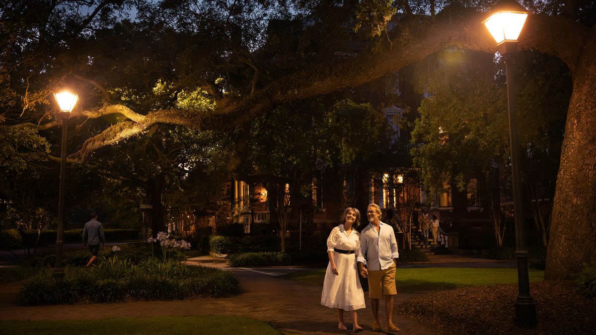 A couple strolls through a Savannah square beneath live oaks and glowing streetlamps at dusk.