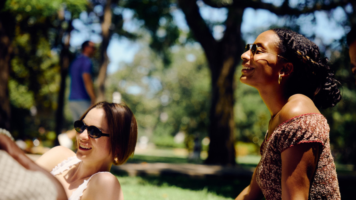 Two friends wearing sunglasses laugh together while relaxing in Savannah’s Forsyth Park.