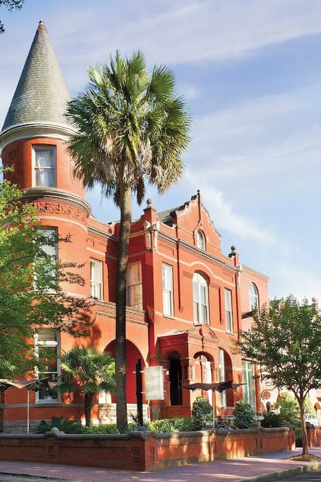 Exterior of Hotel Bardo Savannah, a red-brick historic building with a turreted corner, arched windows, palm trees and a tree-lined street in the foreground.