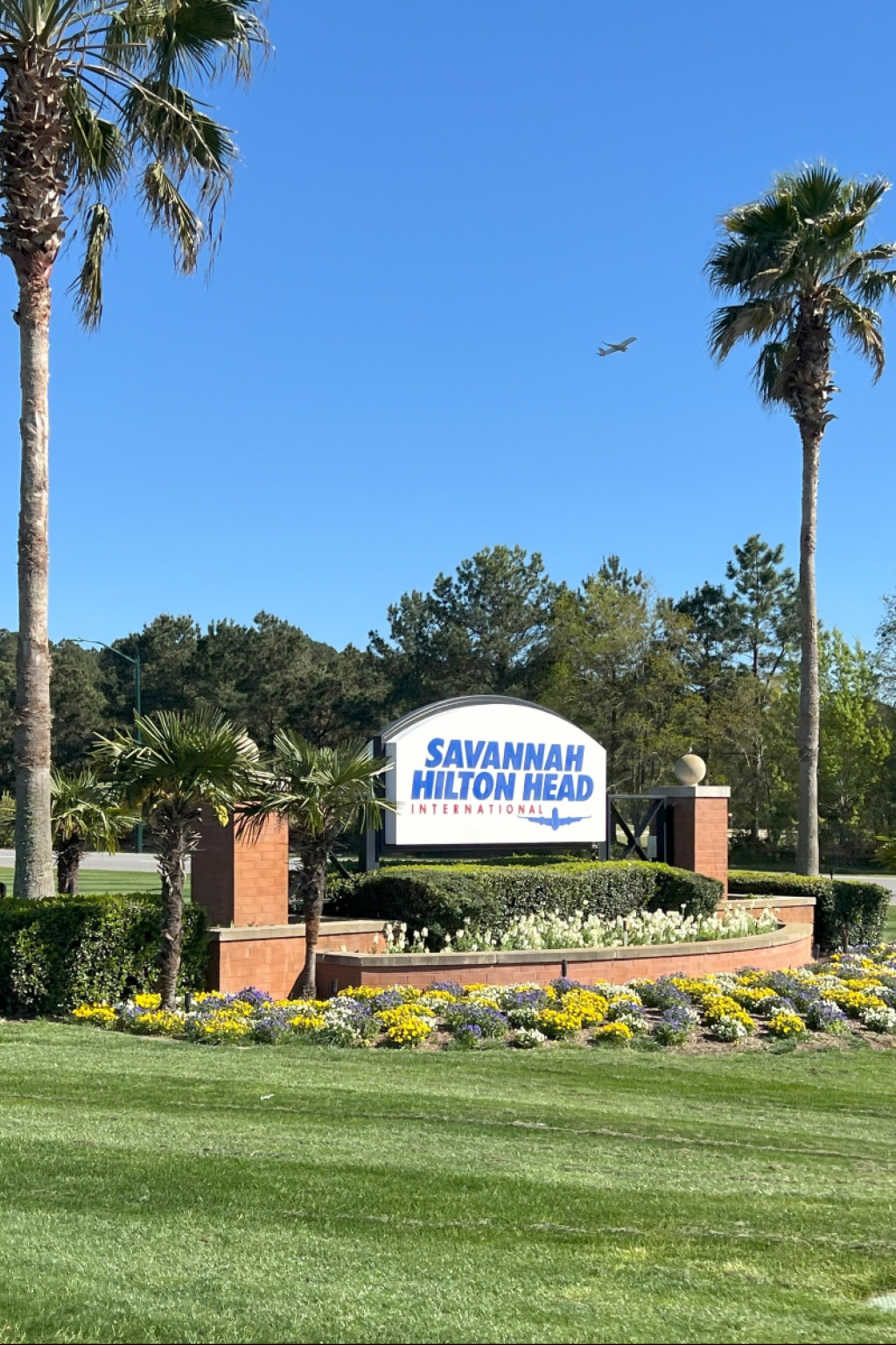 Entrance sign for Savannah Hilton Head International Airport surrounded by palm trees and landscaping under a clear blue sky.