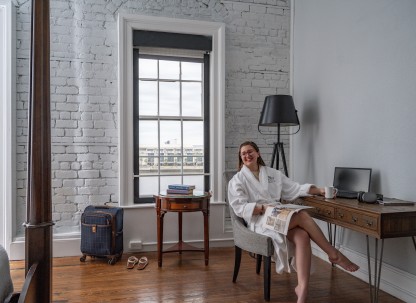 A young woman lounging in a chair in a hotel room wearing a white bathrobe.