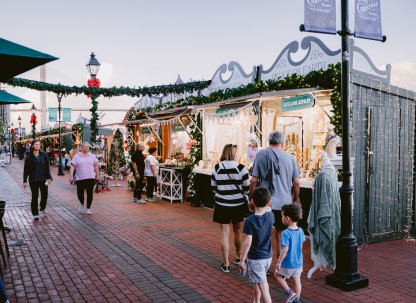 Visitors stroll through the Savannah Christmas Market, lined with festive vendor stalls decorated with garlands, bows, and twinkling lights.