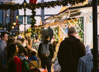 Crowds gather under garlands and twinkling lights at the Savannah Christmas Market at Plant Riverside District, exploring festive vendor stalls decorated for the holidays.