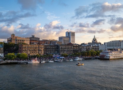 A view of Savannah’s historic River Street at sunset, with boats docked along the riverfront, crowds enjoying the waterfront, and the city skyline—including the gold-domed City Hall—glowing in the evening light.