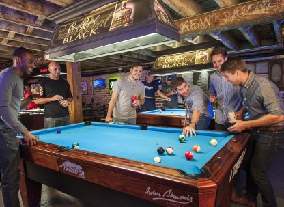 Group of friends playing pool inside a lively Savannah bar, gathered around a blue felt pool table with drinks in hand beneath exposed wooden beams.