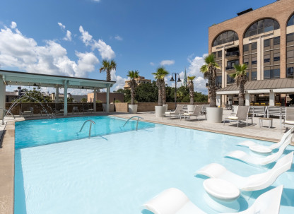 Outdoor pool deck at The DeSoto Savannah featuring a shallow sun shelf with in-water lounge chairs, palm trees, poolside seating and views of the hotel's rooftop terrace beneath a bright blue sky.