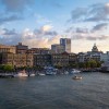 A view of Savannah’s historic River Street at sunset, with boats docked along the riverfront, crowds enjoying the waterfront, and the city skyline—including the gold-domed City Hall—glowing in the evening light.