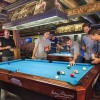 Group of friends playing pool inside a lively Savannah bar, gathered around a blue felt pool table with drinks in hand beneath exposed wooden beams.