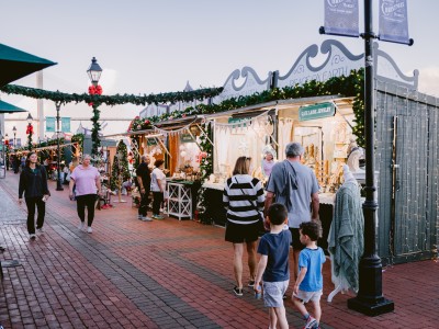 Visitors stroll through the Savannah Christmas Market, lined with festive vendor stalls decorated with garlands, bows, and twinkling lights.
