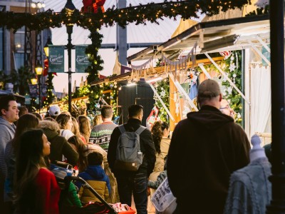 Crowds gather under garlands and twinkling lights at the Savannah Christmas Market at Plant Riverside District, exploring festive vendor stalls decorated for the holidays.