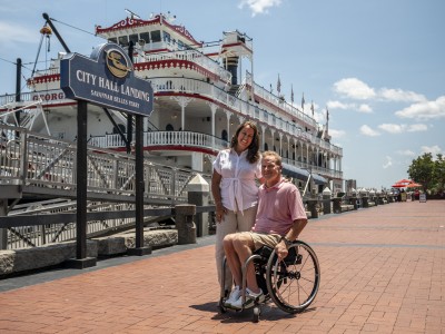 couple on river street in front of riverboat, man in wheelchair