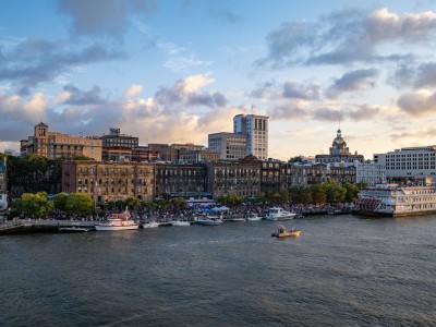 A view of Savannah’s historic River Street at sunset, with boats docked along the riverfront, crowds enjoying the waterfront, and the city skyline—including the gold-domed City Hall—glowing in the evening light.