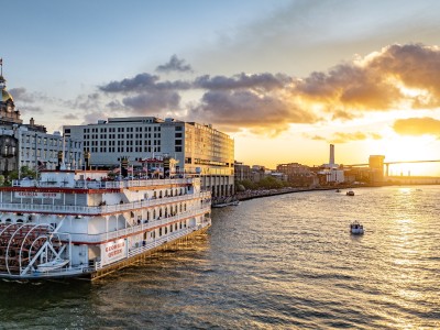The Georgia Queen riverboat cruises along the Savannah River at sunset, with the city skyline and Talmadge Memorial Bridge glowing in the background.