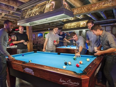 Group of friends playing pool inside a lively Savannah bar, gathered around a blue felt pool table with drinks in hand beneath exposed wooden beams.
